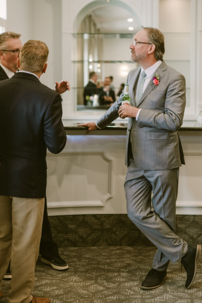 Three men in suits talking at a bar; one man in gray suit holds a drink and leans against the counter.