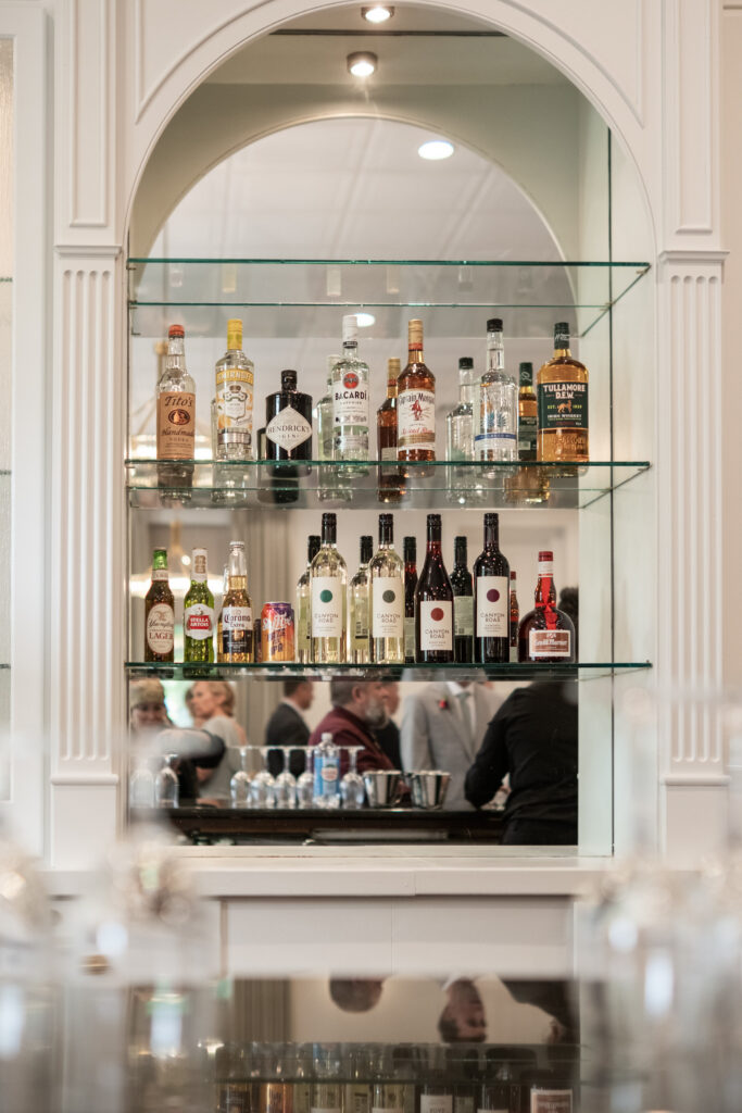 Bottles of liquor displayed on glass shelves behind a bar with people reflected in the mirror.