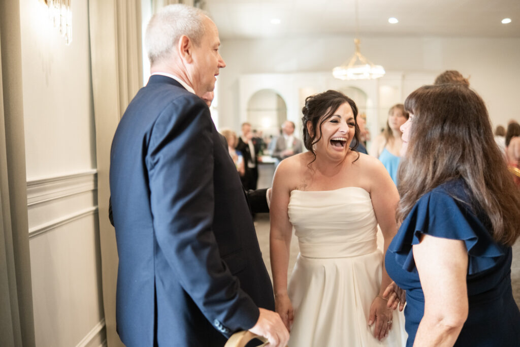 A bride in a white dress laughs joyfully while talking with two guests at a wedding reception.