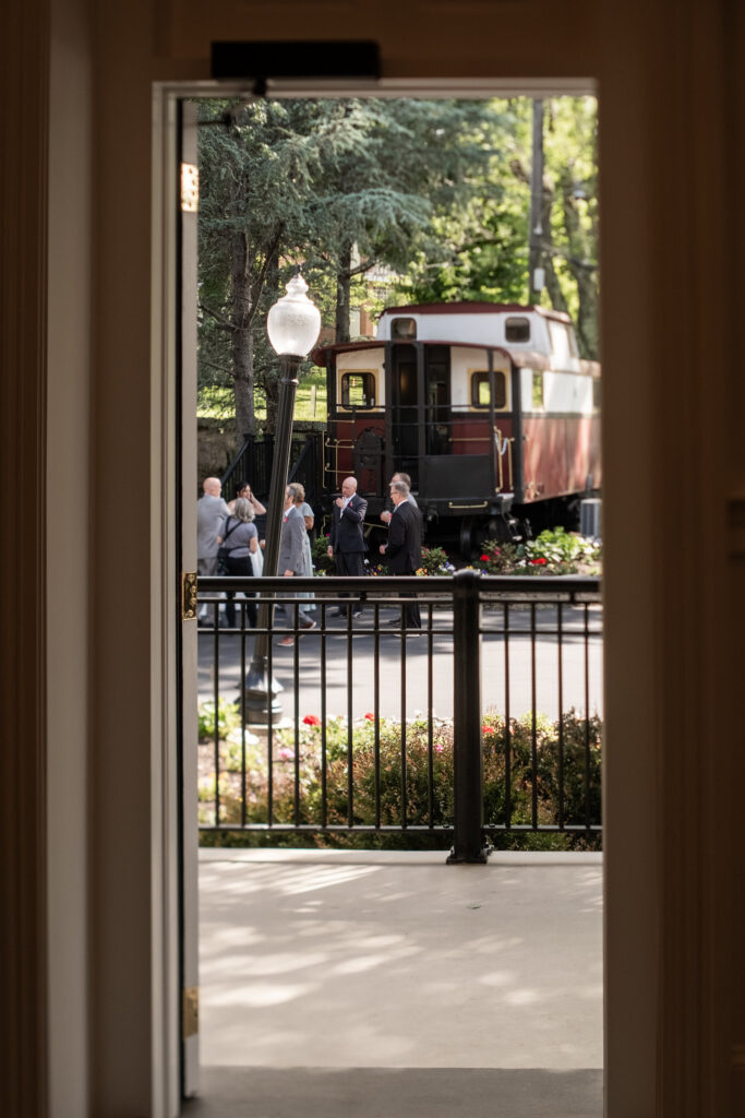 View through an open doorway to people talking near a vintage train car on a tree-lined street.
