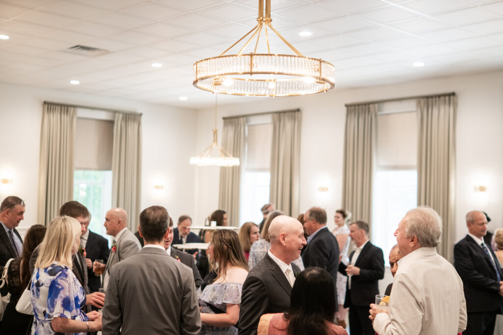 People dressed formally mingle and chat in a bright, elegant room with large windows and chandeliers.