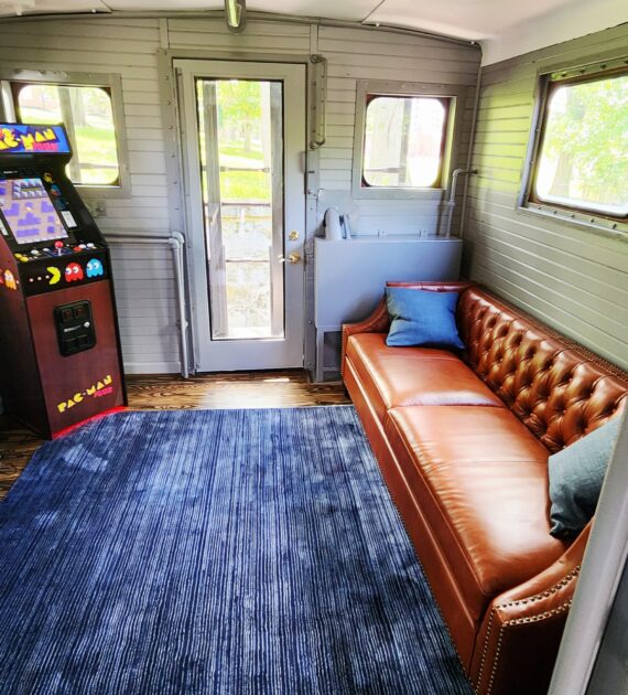 A cozy room with a brown leather couch, blue rug, and a Pac-Man arcade machine by the door.