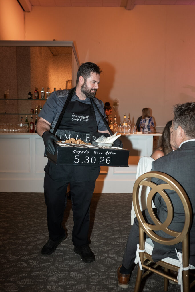 A server in black gloves presents oysters at an event with a bar and seated guests in the background.