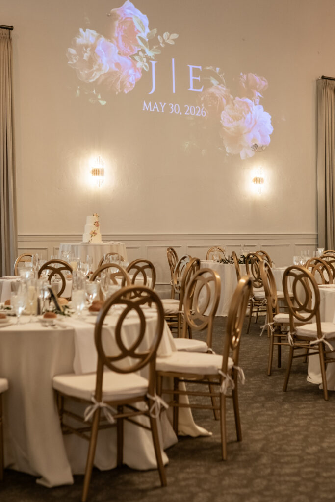 Elegant wedding reception with round tables, gold chairs, and floral date projection on white wall, May 30, 2026.