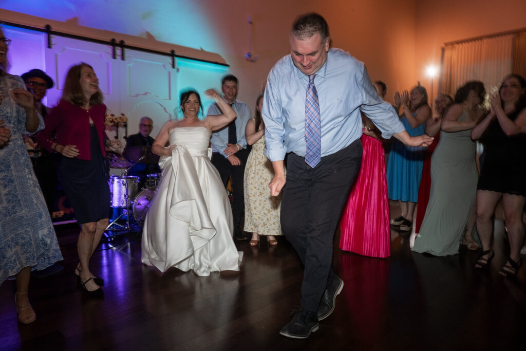 People joyfully dance in a dimly lit room, with a bride smiling among guests on the dance floor.