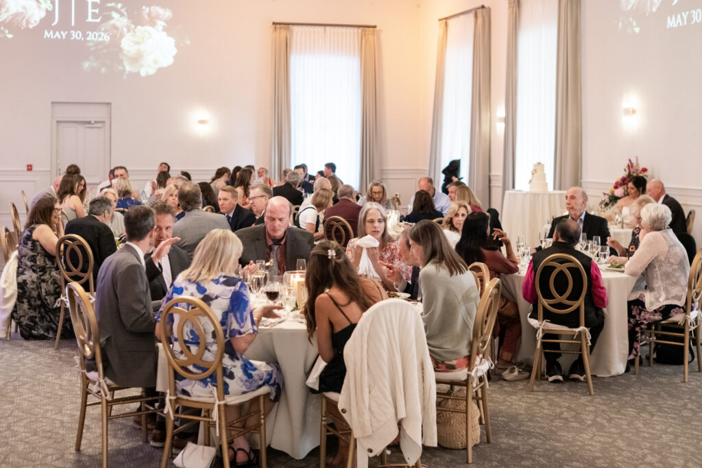 Guests seated at round tables indoors, attending a formal event with white decor and floral accents.