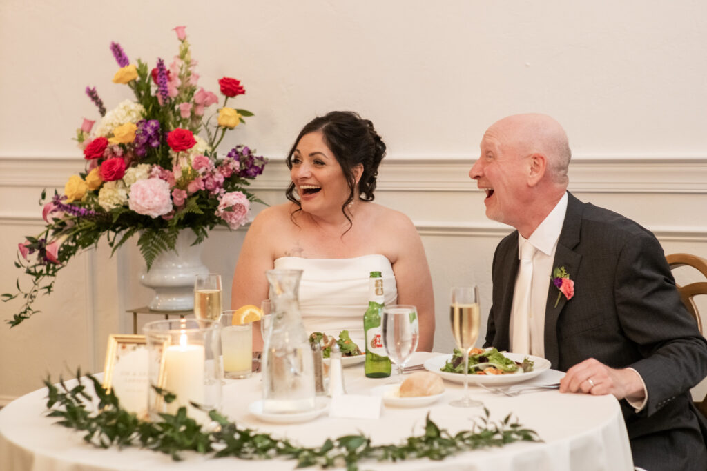 A bride and groom smiling and laughing at a table with flowers, food, and drinks at their wedding reception.