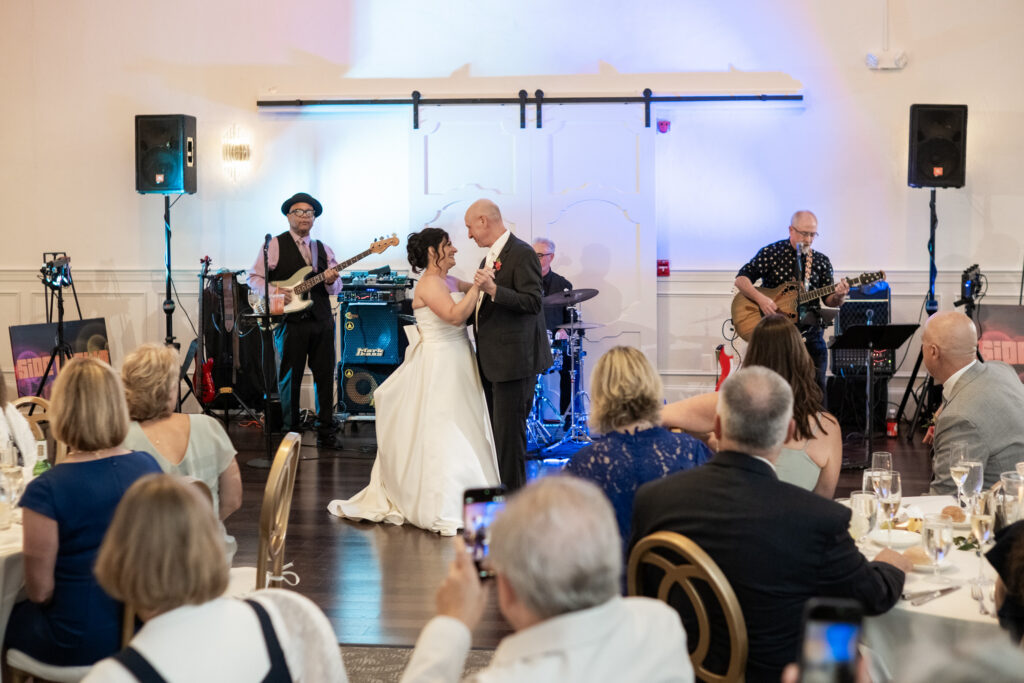 Bride and groom dance while a live band plays, surrounded by guests seated at decorated tables.