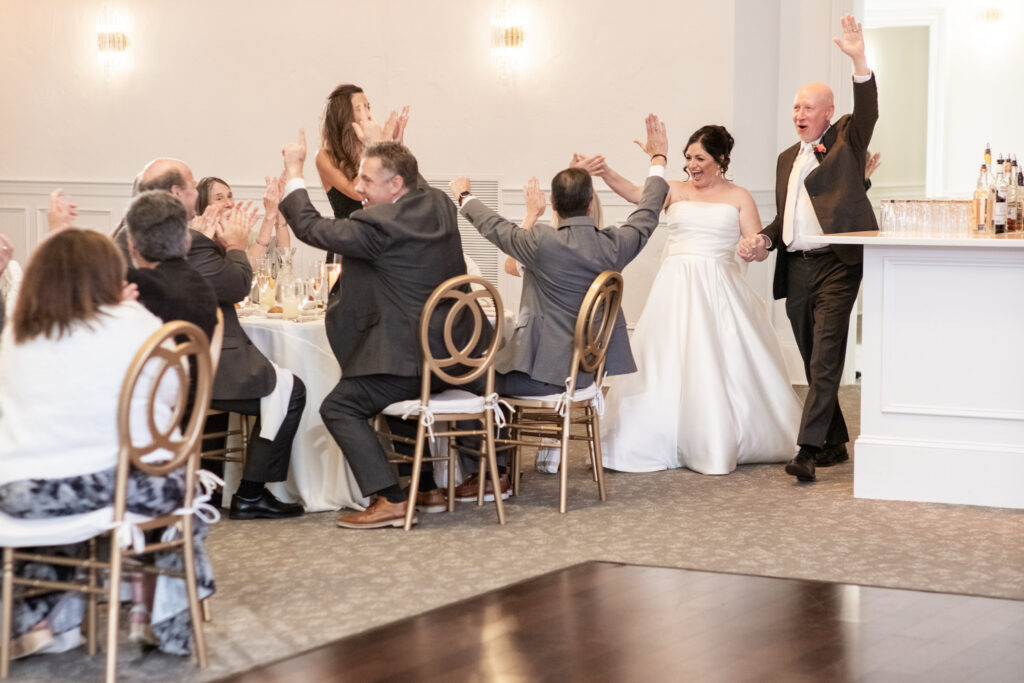 A bride and groom enter a wedding reception, smiling and raising their hands as guests cheer and clap.