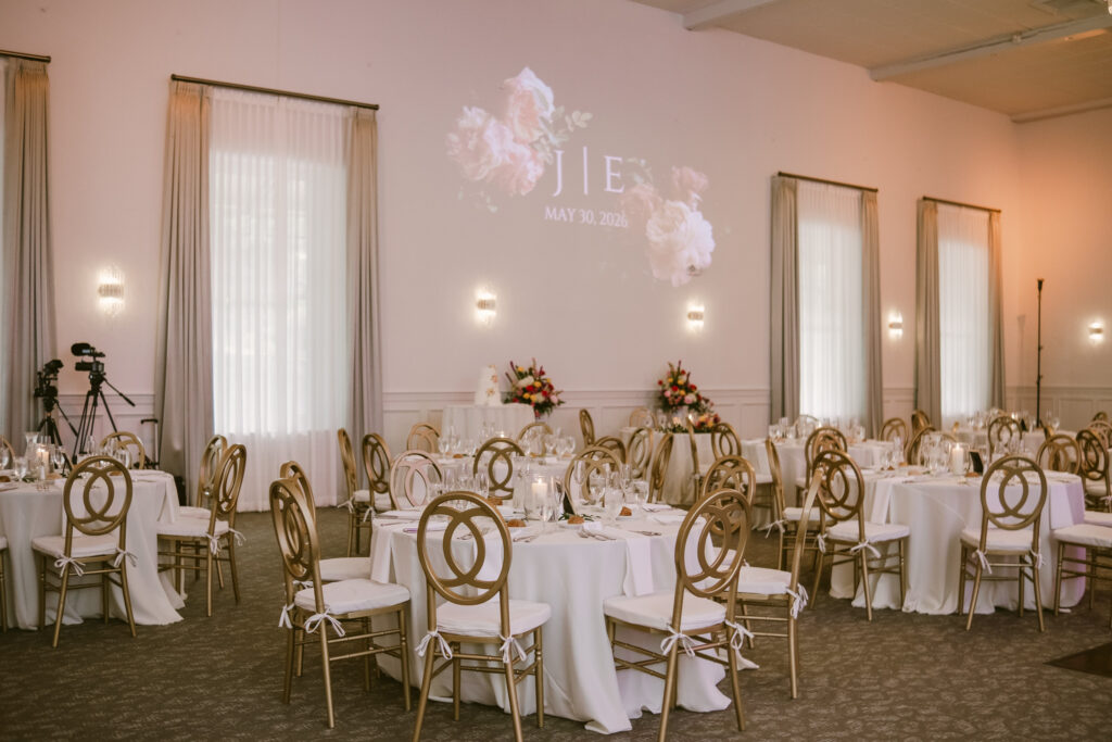 Elegant wedding reception hall with round tables, gold chairs, and floral decorations; "JIE" projected on wall.