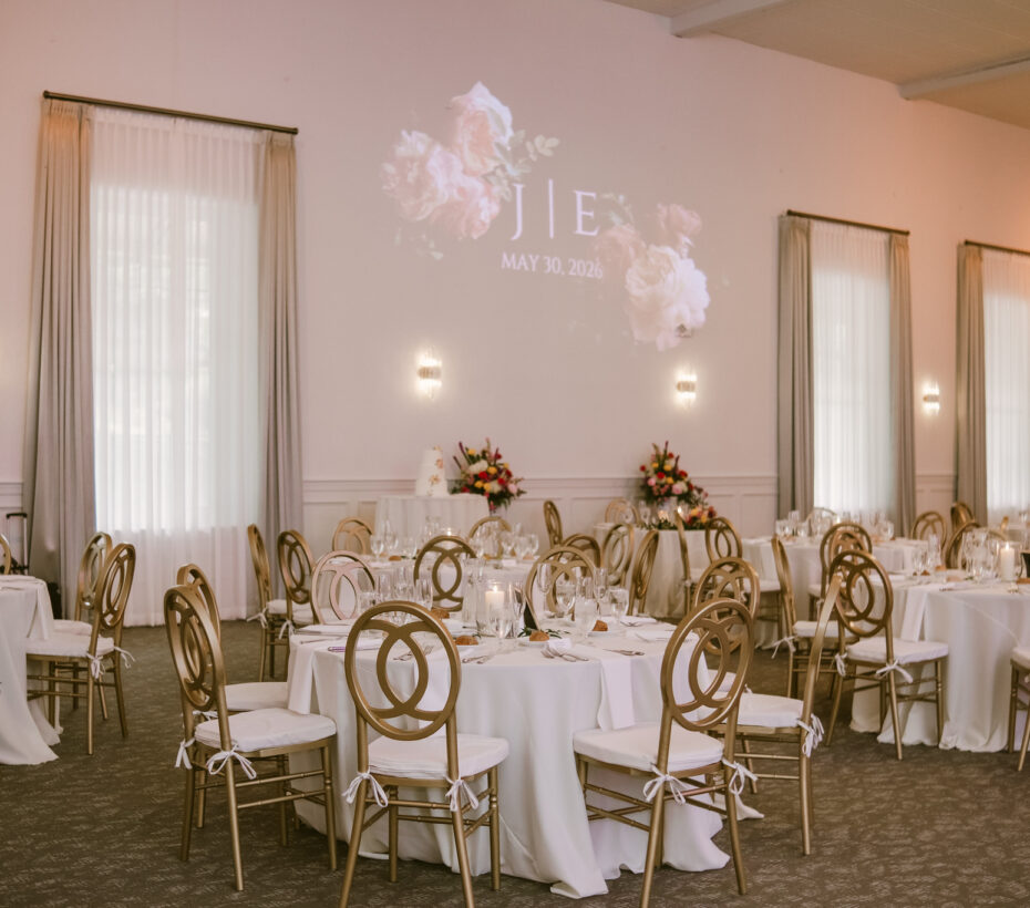 Elegant wedding reception hall with round tables, gold chairs, and floral decorations; "JIE" projected on wall.