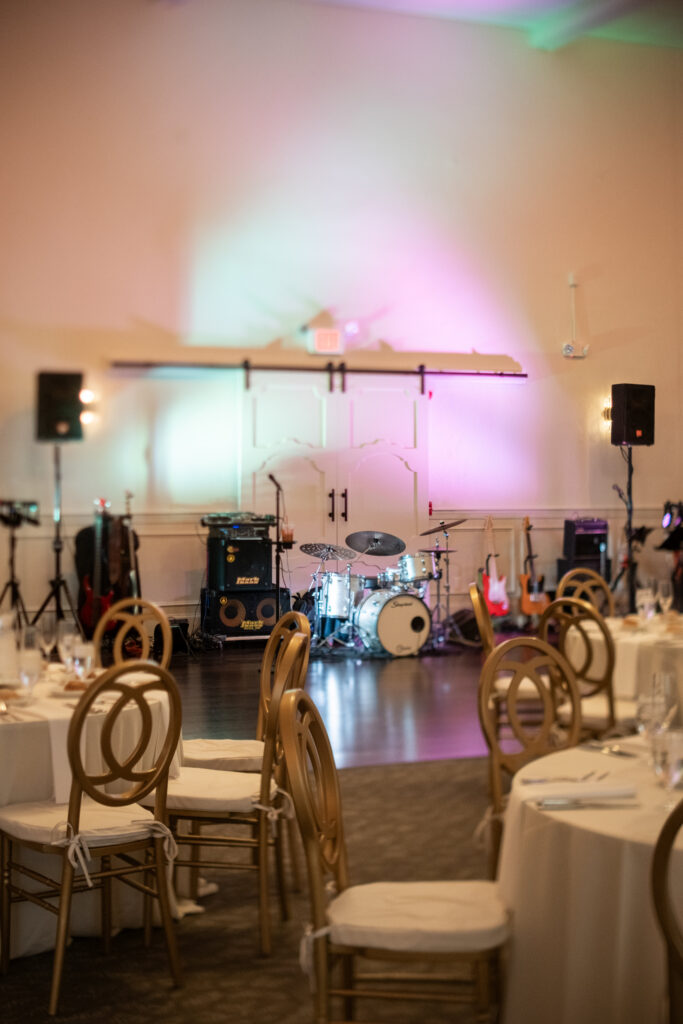 Elegant event space with round tables and chairs, featuring a stage with drums and musical equipment.
