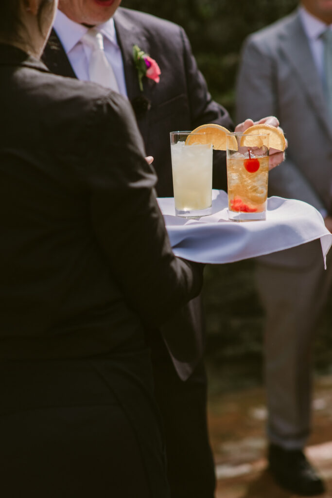A server holds a tray with two cocktails garnished with fruit, as a man takes one drink at an event.