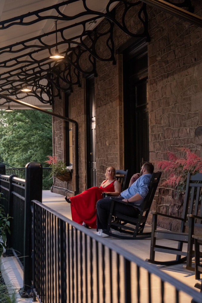 A woman in a red dress and a man sit talking on rocking chairs on a stone building's porch at dusk.
