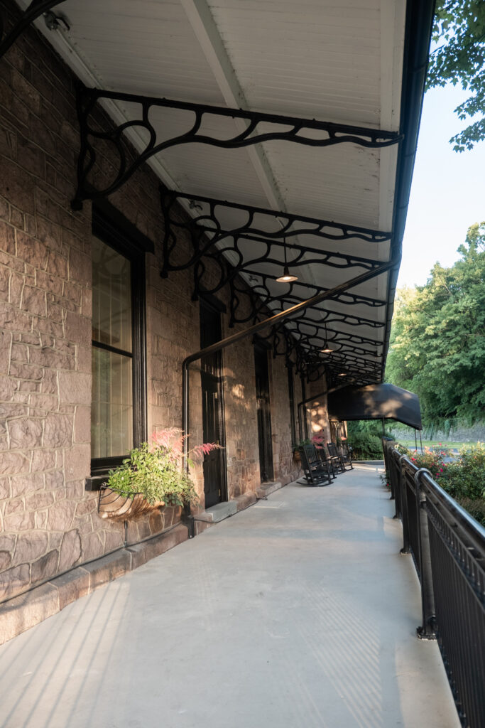 Stone building porch with black iron brackets, rocking chairs, potted plants, and a sloped roof overhead.