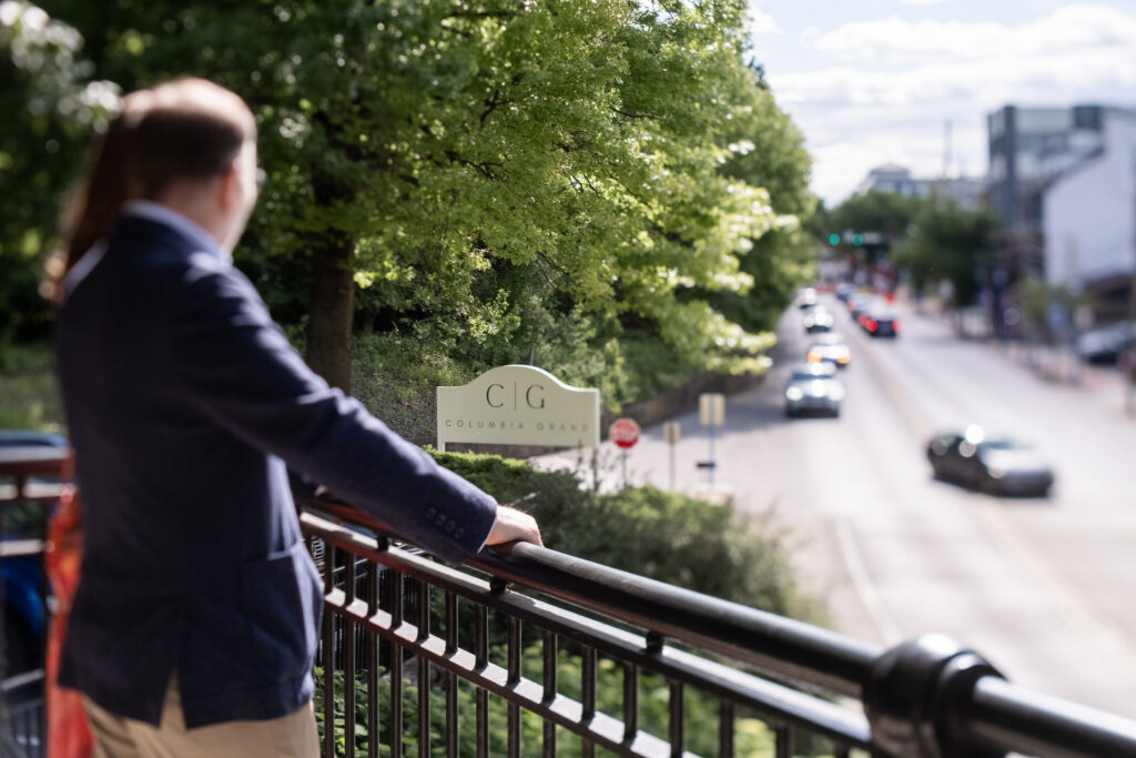 A person in a suit looks out over a railing at a road and a Columbia Group sign among trees.