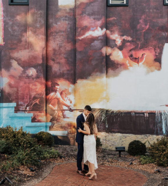 A couple embraces in front of a colorful mural showing industrial and community scenes.