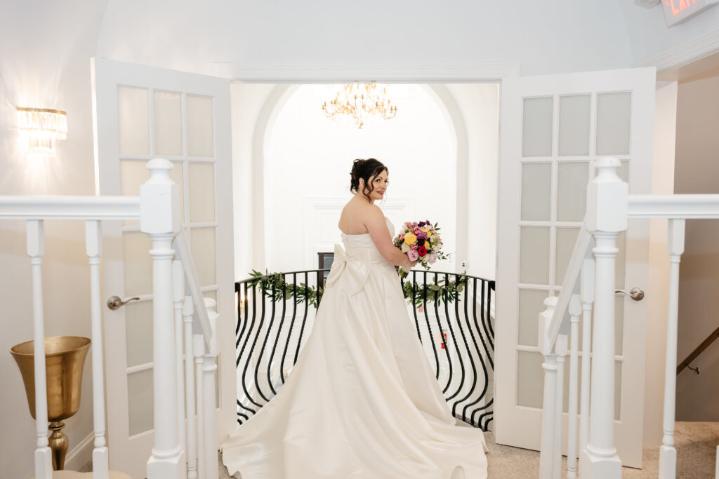 Bride in a white gown holds a bouquet, standing in a bright room with open double doors and a chandelier.