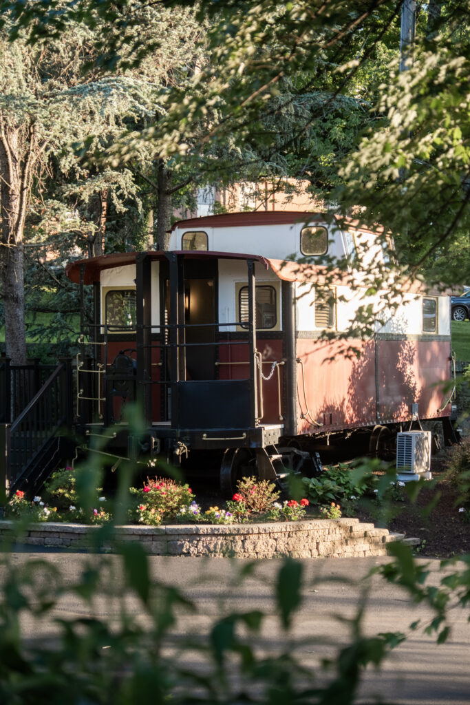 A vintage red train caboose surrounded by trees and flowers in soft sunlight.