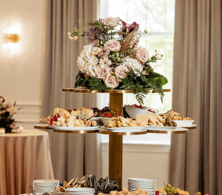 A tiered serving tray with assorted appetizers, cheeses, and meats, topped with a floral arrangement.