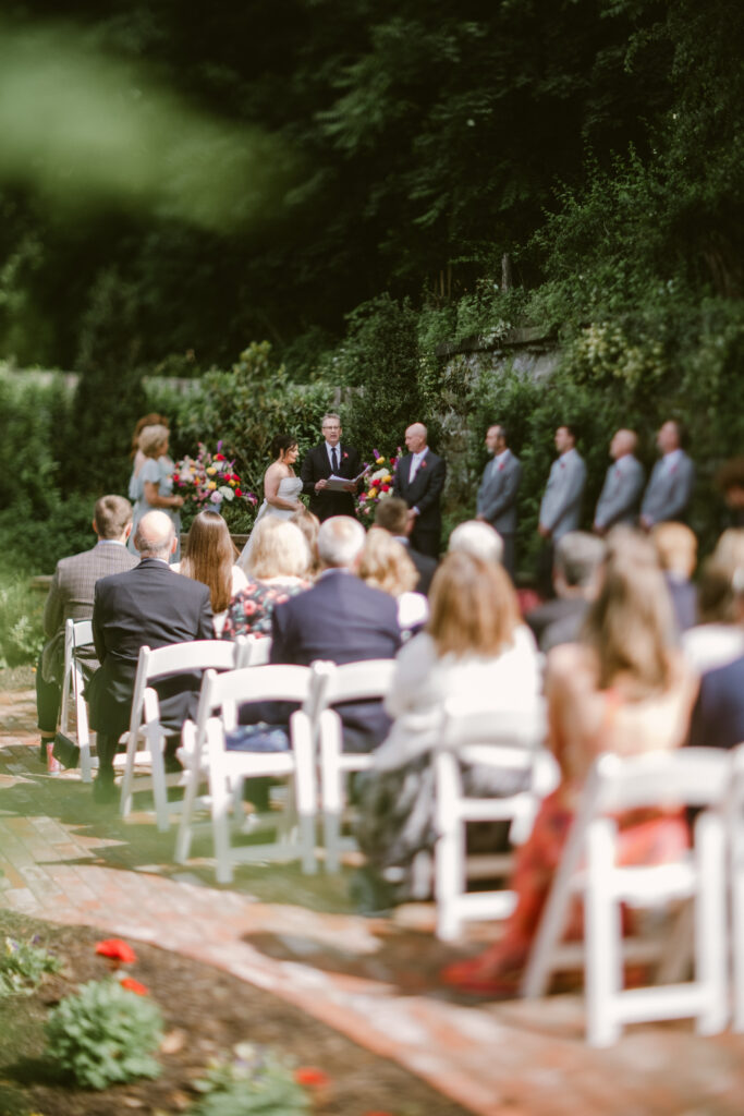 Outdoor wedding ceremony with guests seated, couple standing at the altar, surrounded by greenery.