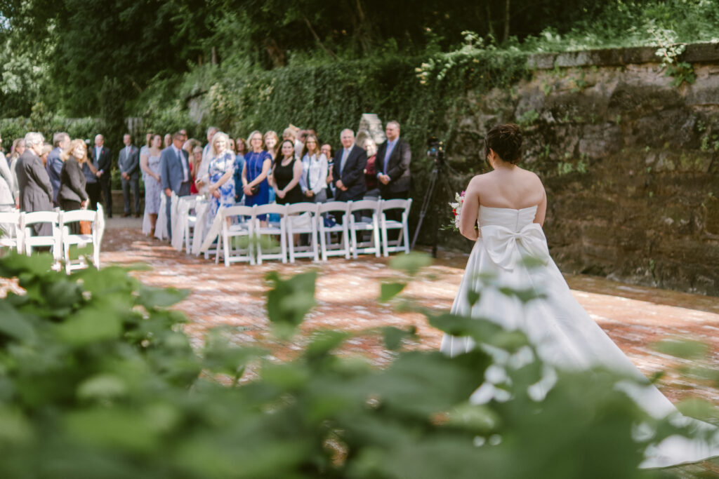 A bride in a white dress walks down an outdoor aisle as guests stand and watch.