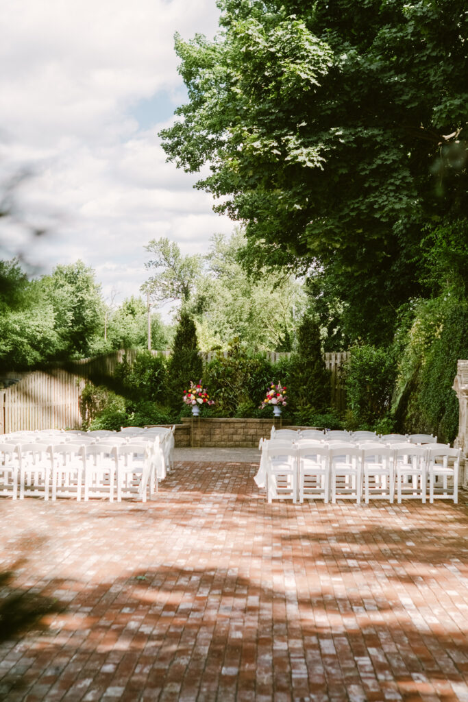 Outdoor wedding ceremony setup with white chairs, brick floor, and floral arrangements under trees.