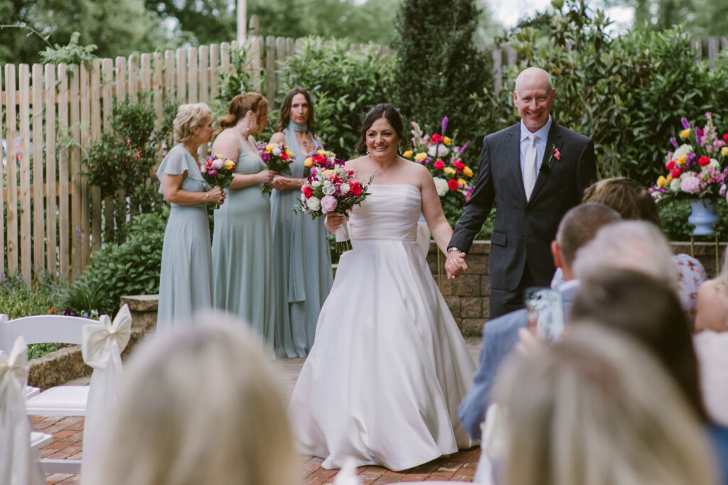 A bride and groom smile, holding hands, walking down the aisle at an outdoor wedding ceremony.