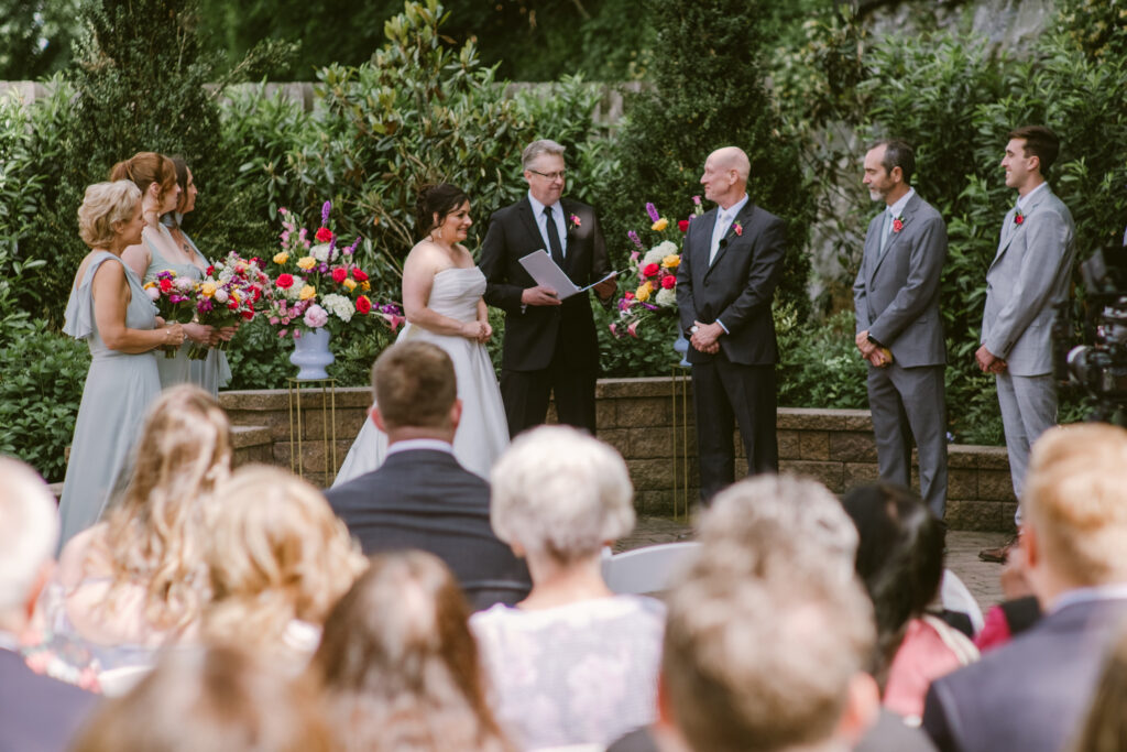 A bride and groom stand with their wedding party during an outdoor ceremony in front of seated guests.
