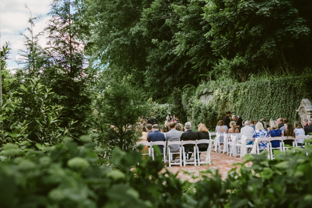 Outdoor wedding ceremony with guests seated on white chairs, surrounded by greenery and trees.