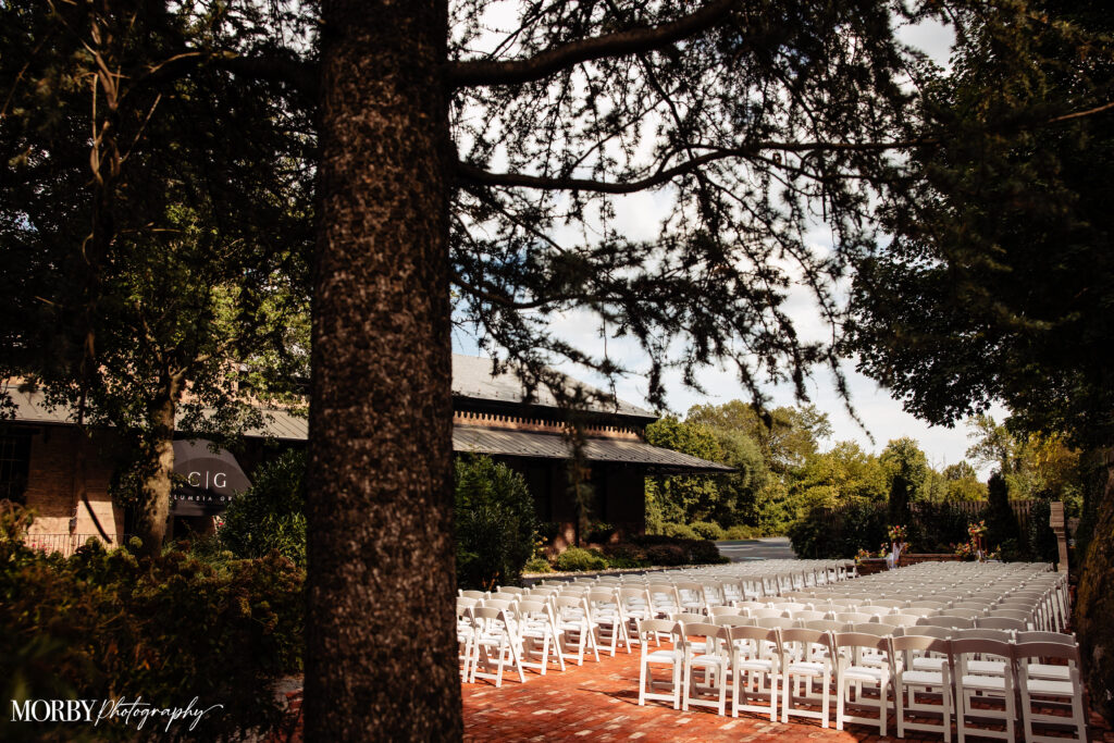 Rows of white chairs set up outdoors on a brick patio for a wedding ceremony, surrounded by trees.