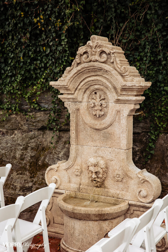 Ornate stone fountain with lion head spout, surrounded by white folding chairs and ivy-covered wall.