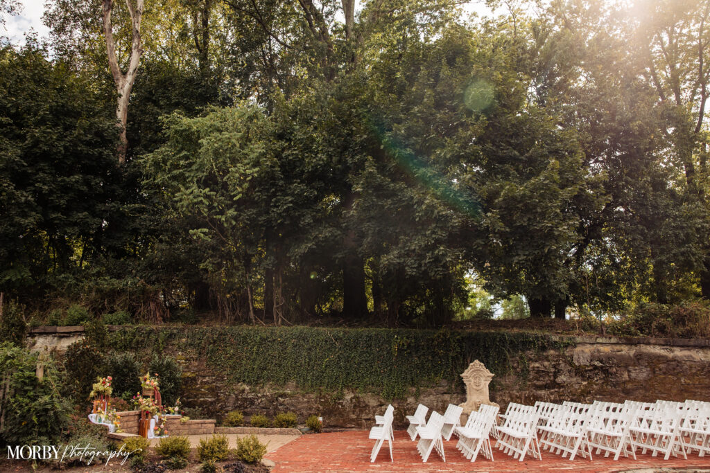Outdoor wedding ceremony setup with rows of white chairs, greenery, and a stone wall in the background.