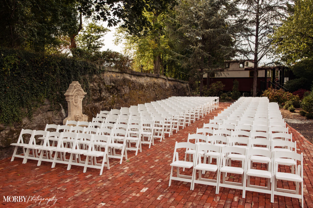 Rows of white chairs set up outdoors on a red brick patio, ready for a wedding ceremony.