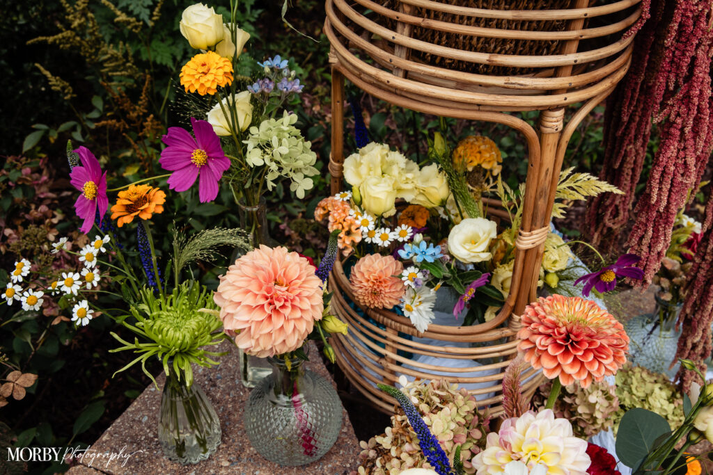 Colorful assorted flowers arranged in glass vases next to a wicker shelf outdoors.