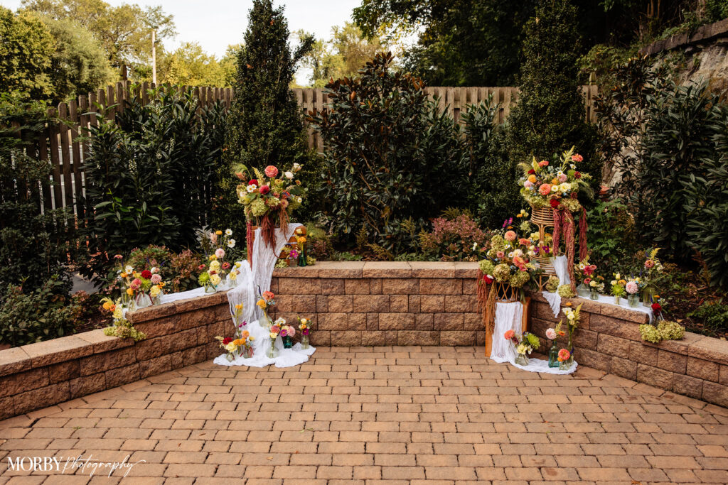 Brick patio with floral arrangements and greenery, set against a stone wall and lush garden background.