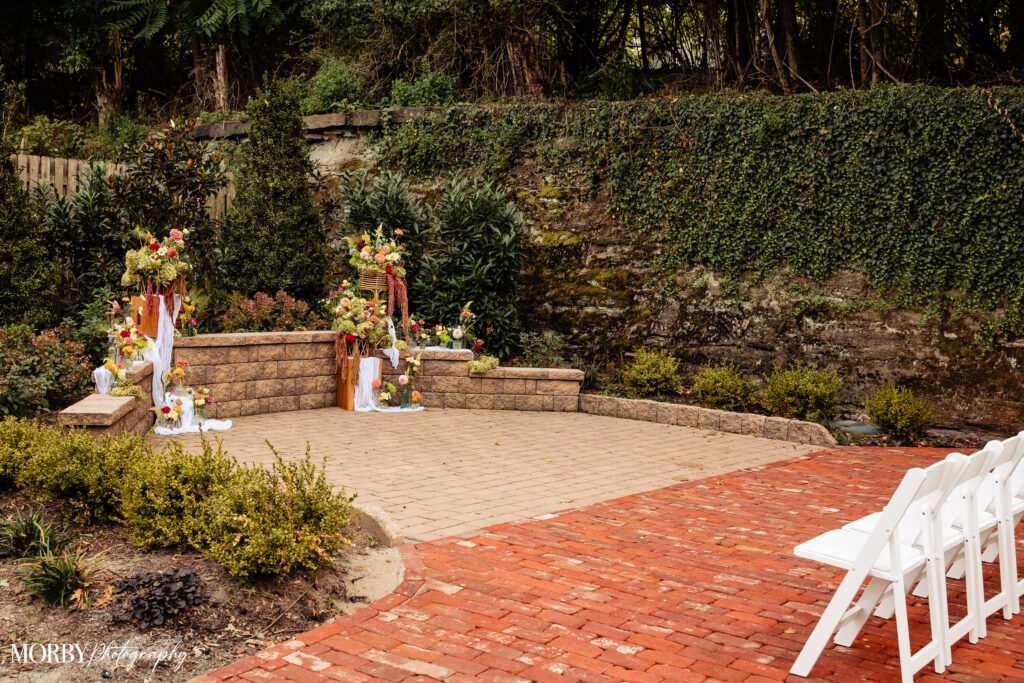 Outdoor wedding altar with floral arrangements and empty white chairs on a red brick patio.