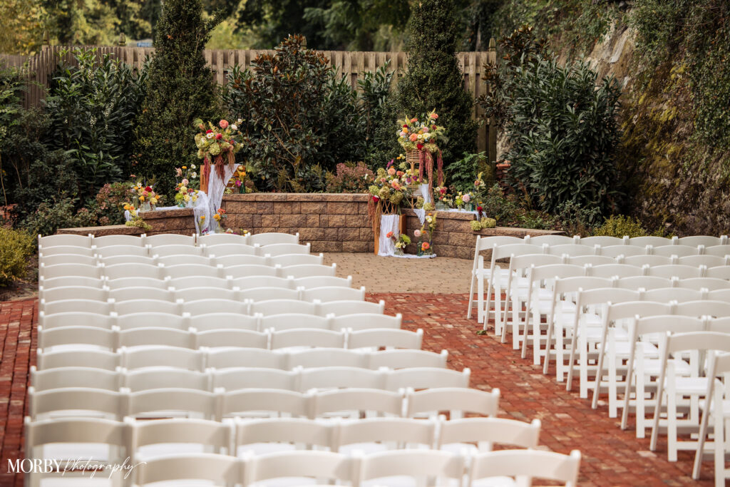 Rows of white chairs face a flower-adorned outdoor wedding altar in a garden setting.