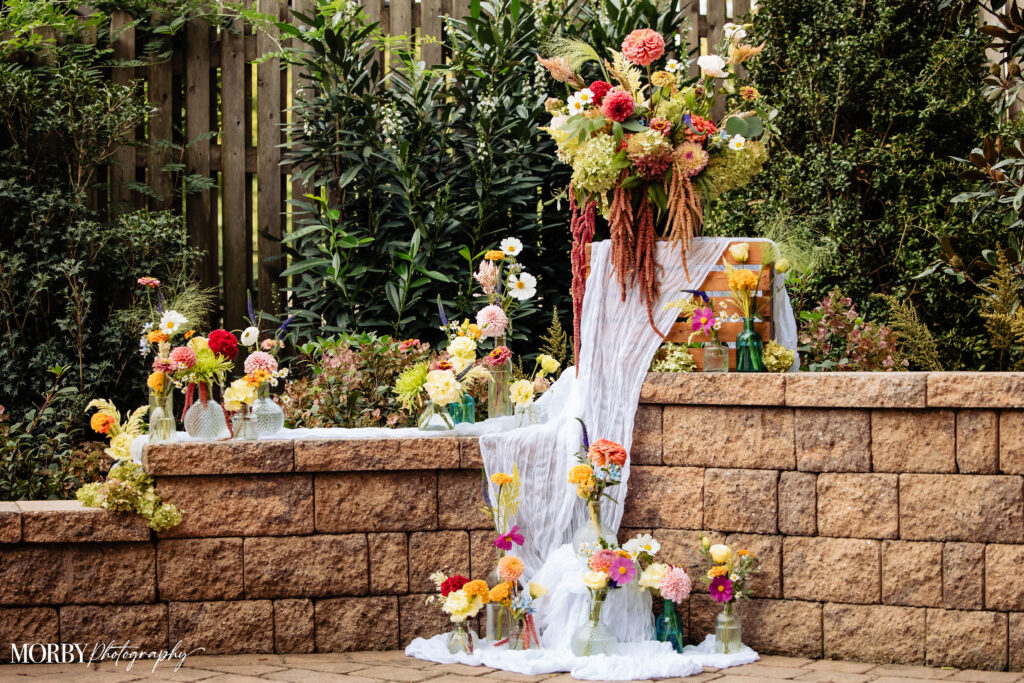 Colorful flowers in glass vases arranged on a brick wall with flowing white cloth, lush greenery behind.