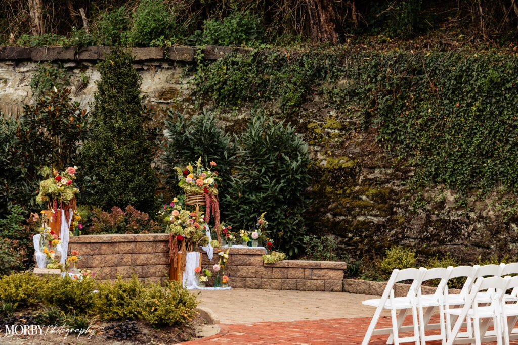 Outdoor wedding ceremony setup with white chairs and floral arrangements in front of a stone wall and greenery.