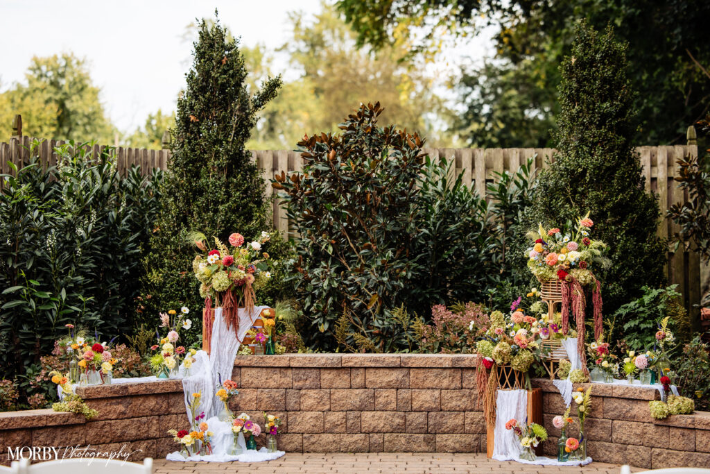 Outdoor wedding altar with colorful flower arrangements and greenery on a stone patio with a wooden fence.