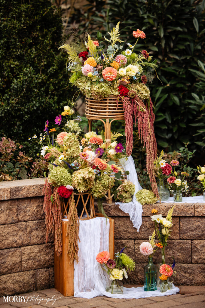 Colorful floral arrangements with various flowers and greenery in vases and baskets against a stone wall.