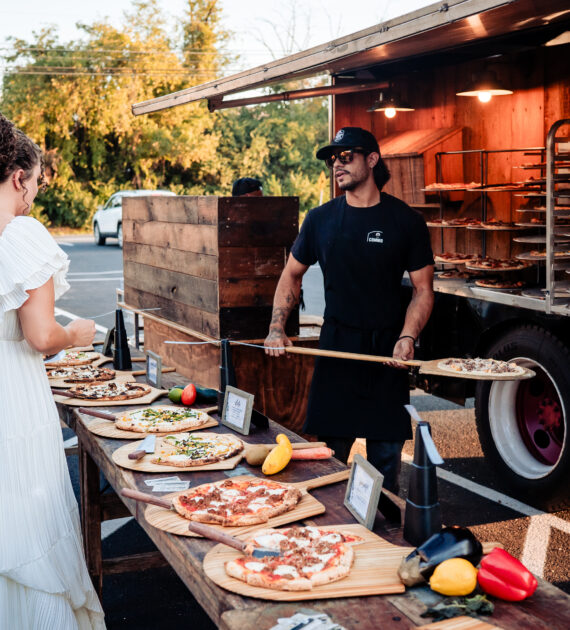 A bride and groom choose pizza from a food truck at an outdoor wedding reception.