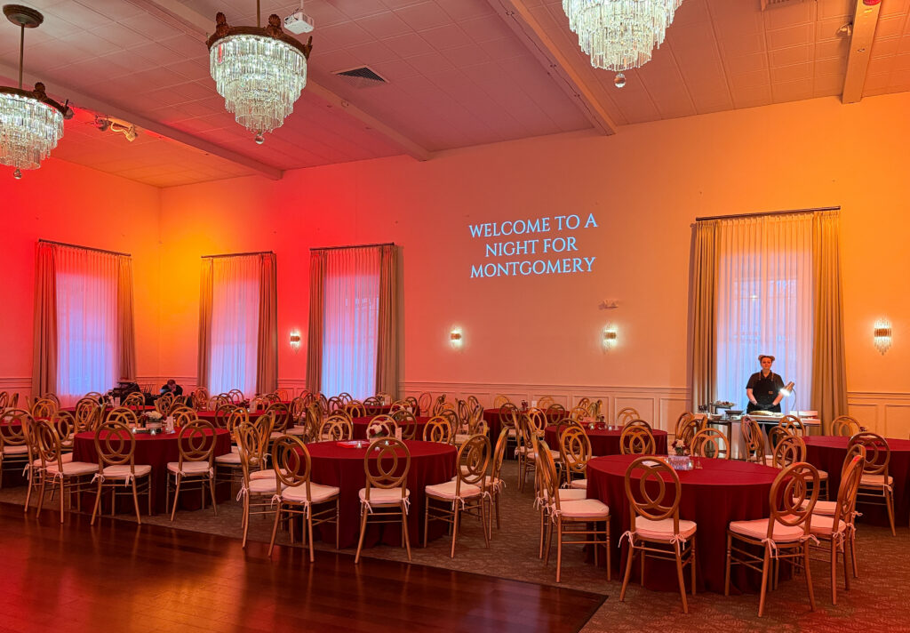 Elegant banquet hall with round tables, red tablecloths, and a wall projection reading "Welcome to a Night for Montgomery.