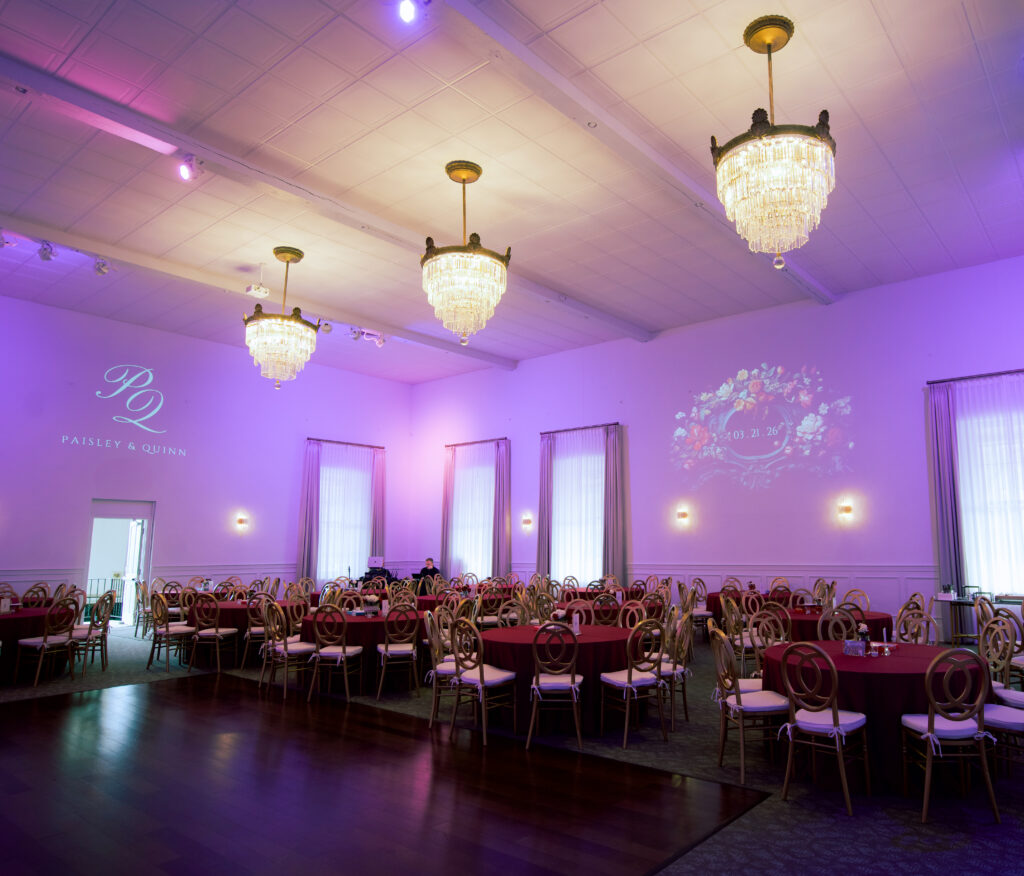 Elegant banquet hall with round tables, gold chairs, chandeliers, and purple ambient lighting.