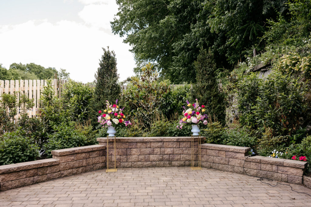 Outdoor stone patio with two tall flower arrangements on stands, surrounded by green bushes and trees.