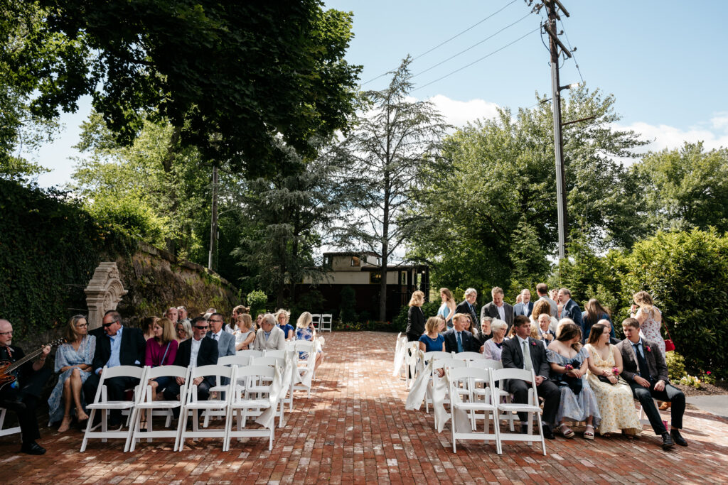 Guests sit on white chairs outdoors at a wedding ceremony, surrounded by trees and greenery under a blue sky.