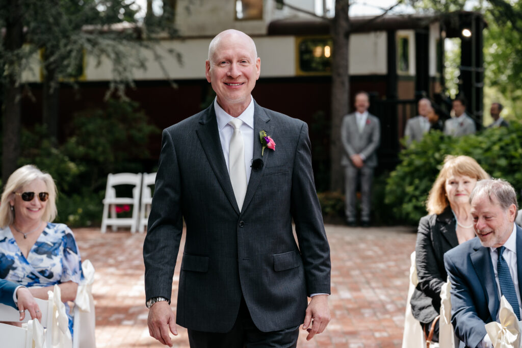 Smiling man in a suit walks down an outdoor aisle, with seated guests watching and trees in the background.