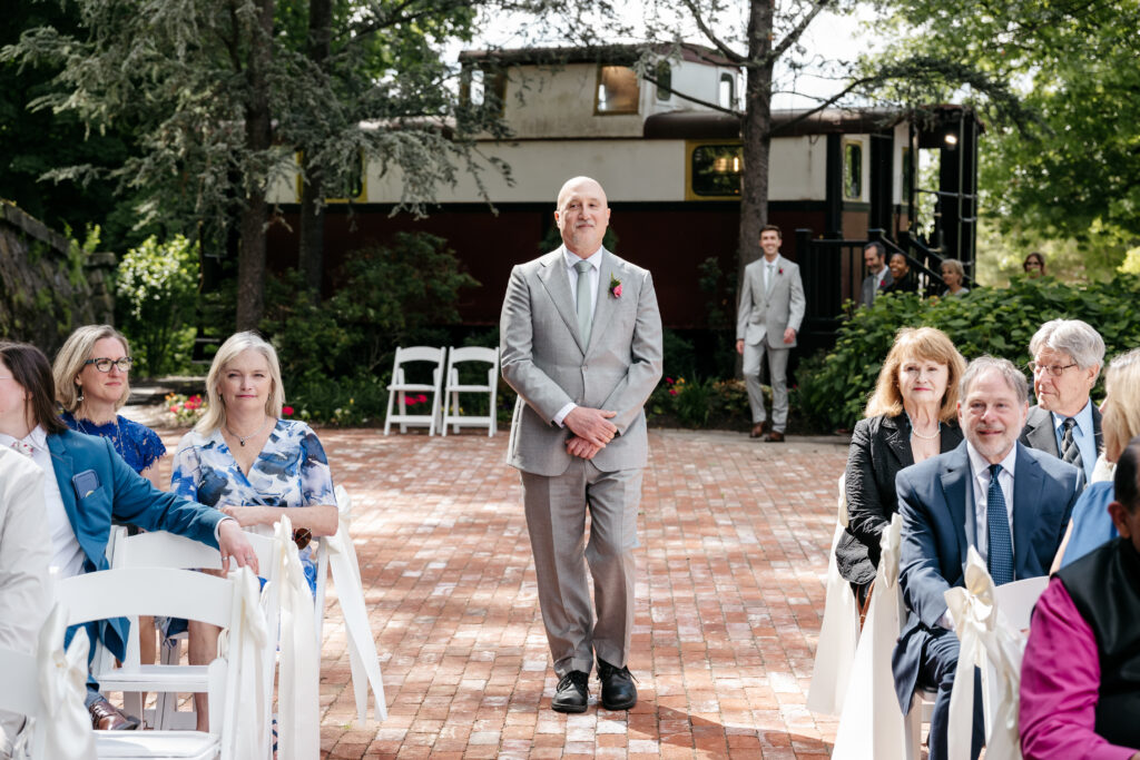 A man in a gray suit stands in an outdoor aisle, surrounded by seated guests at a sunny wedding ceremony.