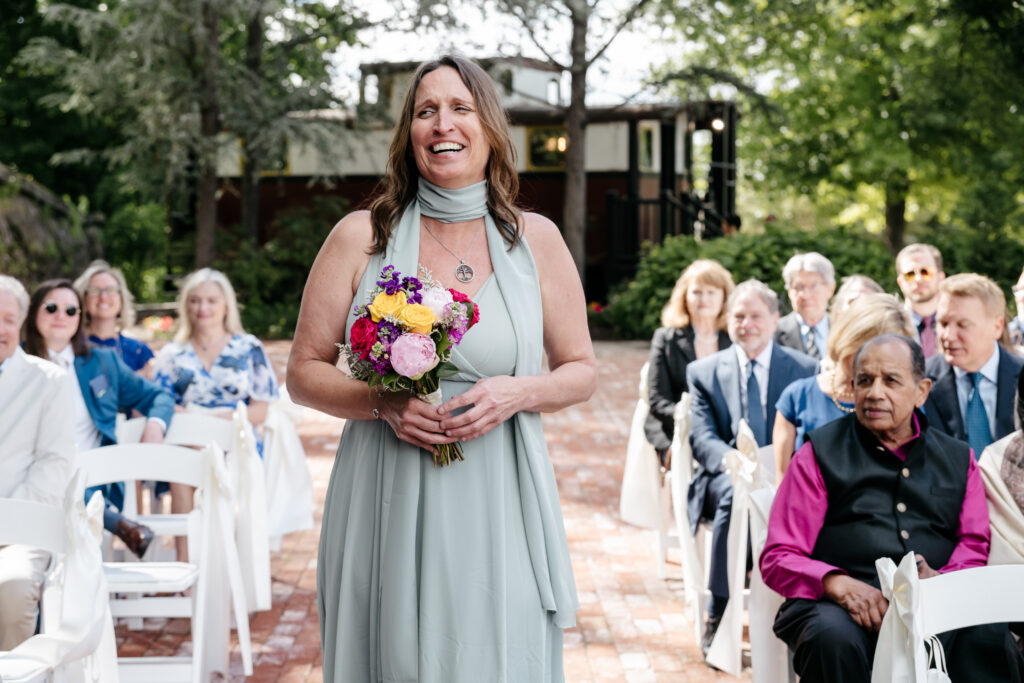 Smiling woman in a light blue dress holds flowers, walking down an outdoor aisle with seated guests watching.